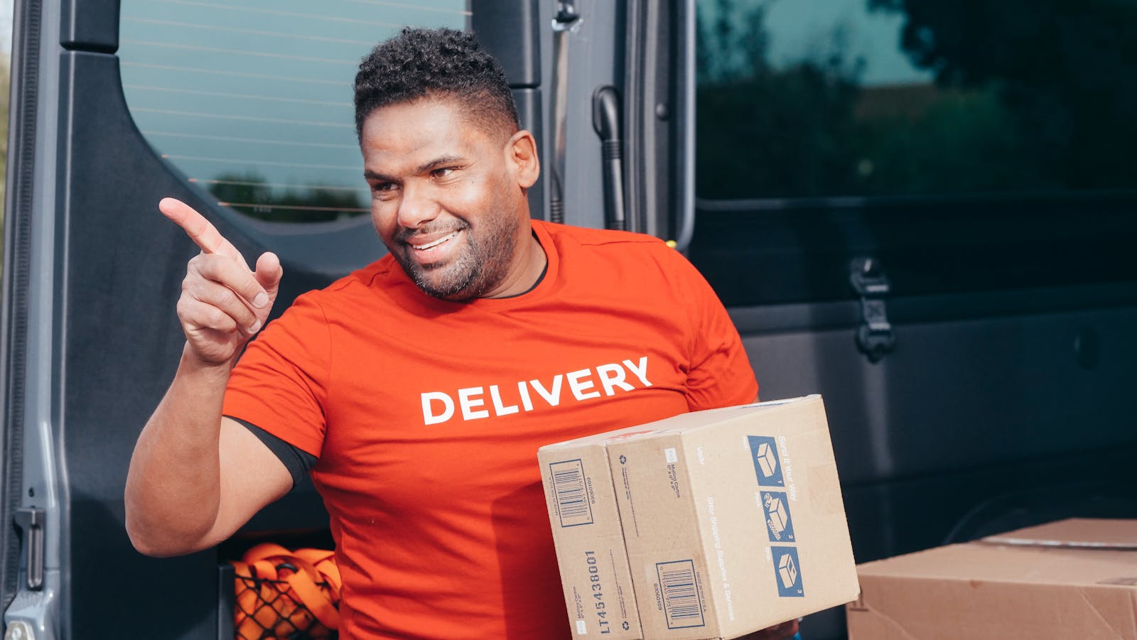 Friendly delivery worker in red shirt holding a package and pointing outdoors.